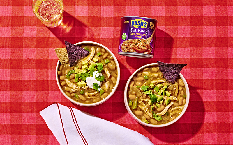 Two bowls of white chicken chili next to a can of Bush's Slow-Simmered White Chili Magic on a red tablecloth